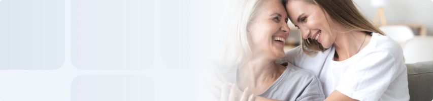 Jüngere Frau umarmt lächelnd eine ältere Frau – beide lehnen die Stirn aneinander.